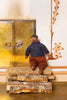 A small knitted teddy bear doll wearing a blue patterned sweater, brown pants, and a green hat, sitting on a stack of birch logs in front of a rustic wooden door and decorative wall.