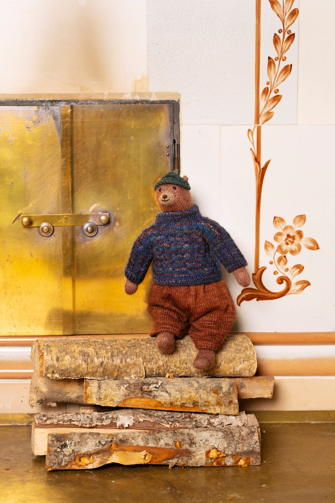 A small knitted teddy bear doll wearing a blue patterned sweater, brown pants, and a green hat, sitting on a stack of birch logs in front of a rustic wooden door and decorative wall.