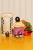 A handmade doll figurine with a black knit head, yellow striped sweater, and red-and-white striped skirt, standing on a yellow tabletop next to a wooden memory board and a red pot of vegetables.