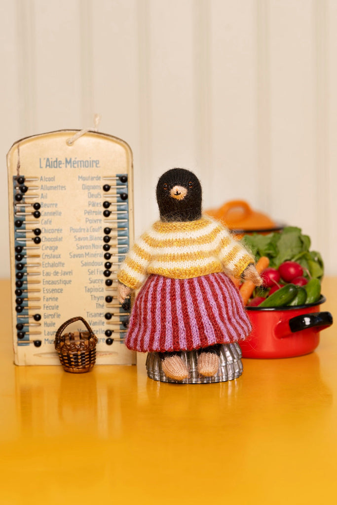 A handmade doll figurine with a black knit head, yellow striped sweater, and red-and-white striped skirt, standing on a yellow tabletop next to a wooden memory board and a red pot of vegetables.
