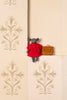 A small plush gray bear wearing a bright red dress sits on a cream-colored tiled ledge next to a small brown handbag. The background features elegant beige damask patterned tiles.