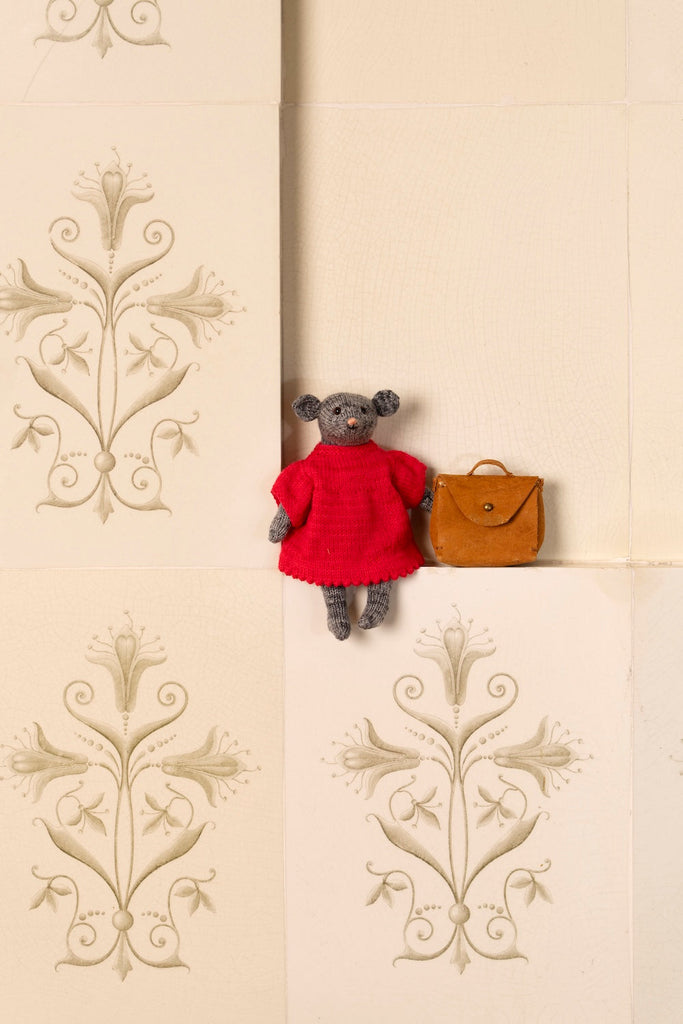 A small plush gray bear wearing a bright red dress sits on a cream-colored tiled ledge next to a small brown handbag. The background features elegant beige damask patterned tiles.