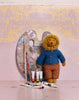 A knitted lion doll with a golden mane stands in front of a paint palette and brushes, with several paint tubes on the surface in front. The doll is wearing a blue cardigan and brown patterned pants against a textured pastel background.