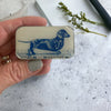 Hand holding a small rectangular metal box with a blue dog illustration on a speckled surface.