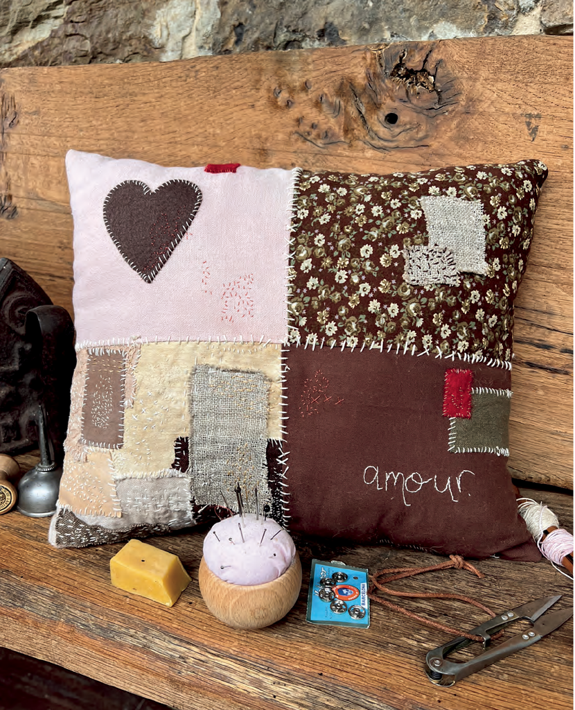 A square patchwork pillow with a rustic handmade aesthetic, featuring stitched house shapes, floral fabric, and the word 'amour' on one side, displayed on a wooden bench with sewing tools.