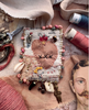 A handmade embroidered fabric pouch or bag with a stitched heart motif, beads, shells, thread, and small charms, arranged on a textile surface with scissors, spools of thread, and fabric around it.