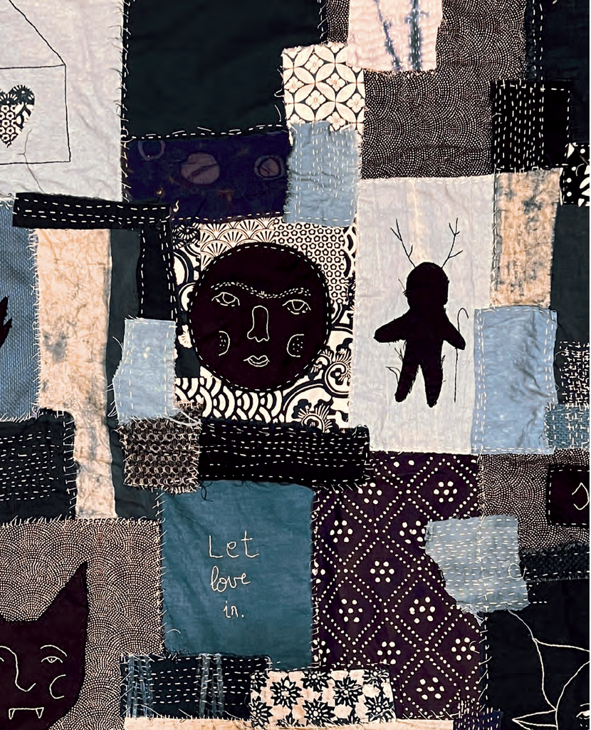 A close-up of a patchwork quilt featuring various fabric squares in dark blue, beige, black, and white, decorated with silhouettes, patterns, and the handwritten text 'Let love in.'.