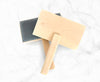 Two wooden brushes with light natural wood handles and rectangular heads, one with a dark gray textured bristle pad and one with a smooth pale wood pad, arranged on a white marble surface.