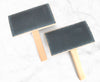 Two brushes with dark gray textured bristles and light natural wood handles, arranged diagonally on a white marble surface.