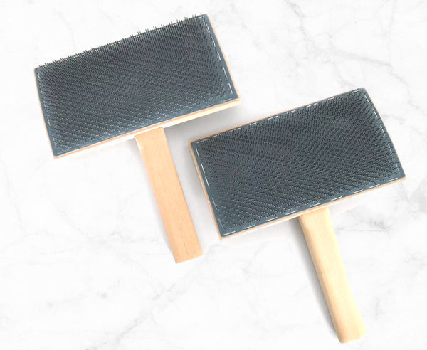 Two brushes with dark gray textured bristles and light natural wood handles, arranged diagonally on a white marble surface.