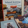 A book titled "Island Knitting" is shown standing upright, with other knitting-related books partially visible in front. The cover features a stack of knitted garments in earthy colors and the subtitle "Beautiful Knitting Patterns Inspired by Coastal Landscapes.