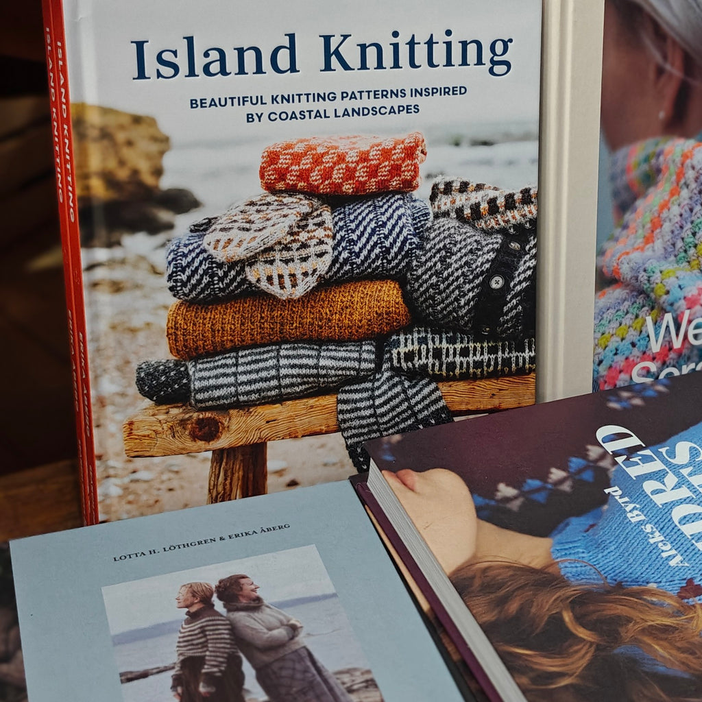 A book titled "Island Knitting" is shown standing upright, with other knitting-related books partially visible in front. The cover features a stack of knitted garments in earthy colors and the subtitle "Beautiful Knitting Patterns Inspired by Coastal Landscapes.