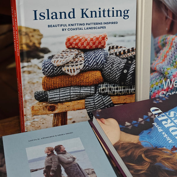 A book titled "Island Knitting" is shown standing upright, with other knitting-related books partially visible in front. The cover features a stack of knitted garments in earthy colors and the subtitle "Beautiful Knitting Patterns Inspired by Coastal Landscapes.