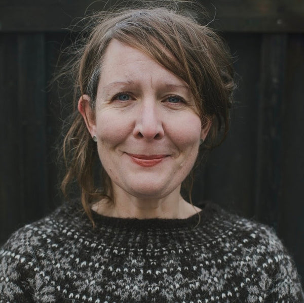 A close-up portrait of a woman with brown hair wearing a black and white patterned knit sweater, set against a dark wooden background.