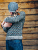 A man wearing a gray knit sweater, blue jeans, and a gray flat cap holds a small brown and white dog against his chest, standing in front of a rustic wooden plank wall.