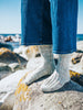 A close-up of a person standing on rocks by the sea, wearing blue jeans and light gray ribbed knit socks.