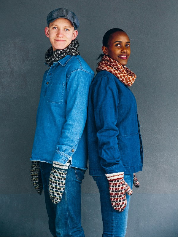 Two models standing back-to-back wearing matching blue outerwear, blue jeans, patterned knit scarves, and knit mittens against a dark gray studio background.
