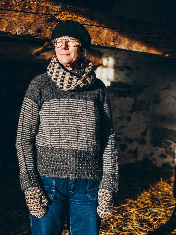 A person wearing a dark gray and beige patterned knit sweater, a matching patterned scarf, a black beret, and knit gloves, standing in a rustic indoor setting with warm light.