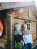 Two people standing outside a rustic wooden cabin, with one wearing a dark patterned sweater and green pants, and the other wearing a gray and white color-blocked sweater and blue pants.