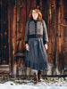 A woman stands outdoors in front of a rustic wooden wall, wearing a dark gray knit cardigan over a black top, a long dark gray skirt, and brown boots with snow visible on the ground.