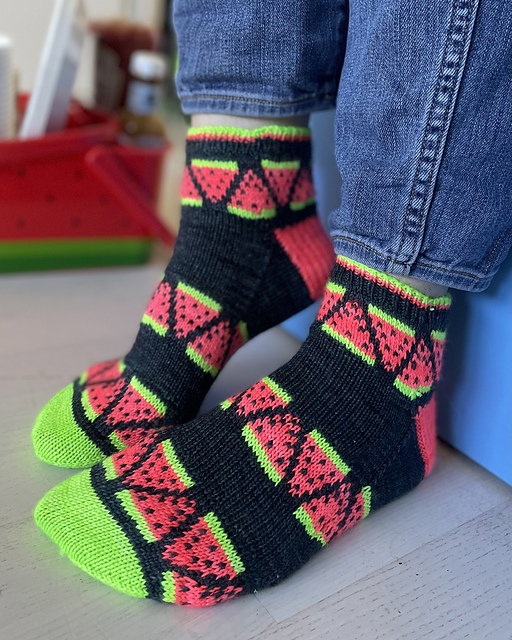 Person wearing black socks with red and green watermelon pattern on a light wooden floor.
