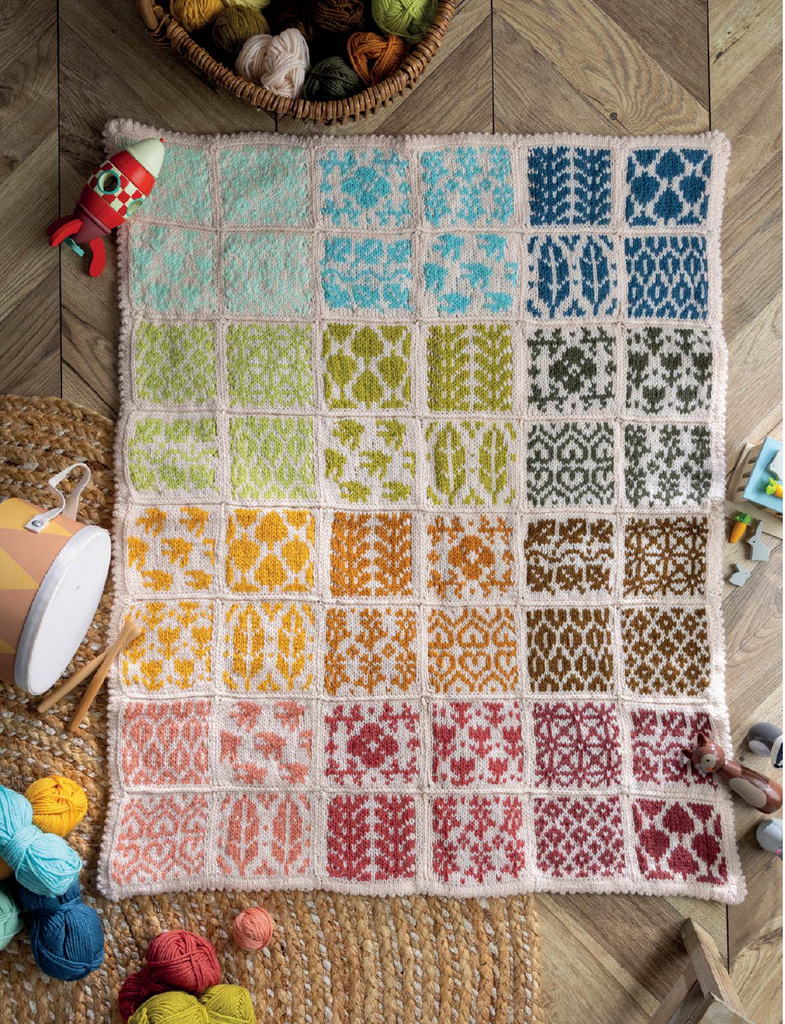 A handmade crocheted blanket laid flat on a wooden floor, featuring a grid of colorful patterned squares in pastel and bright tones. The blanket is surrounded by knitting props including yarn balls, a wooden mallet, and a basket of yarn.