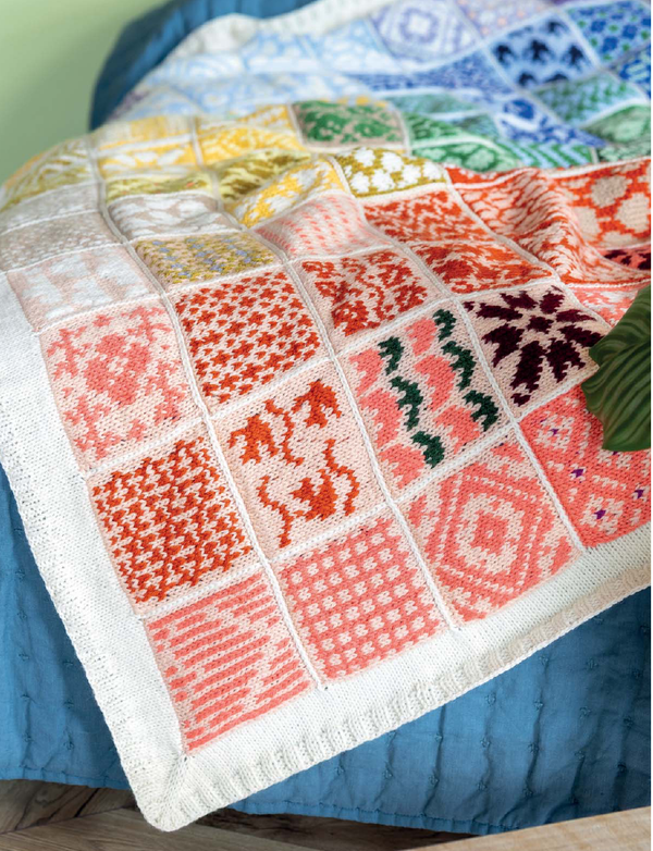 A close-up of a knitted patchwork blanket draped over a blue quilted bed. The blanket features multiple square panels with various colorful knit patterns in orange, red, pink, yellow, green, and blue on an off-white base.