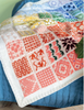 A close-up of a knitted patchwork blanket draped over a blue quilted bed. The blanket features multiple square panels with various colorful knit patterns in orange, red, pink, yellow, green, and blue on an off-white base.