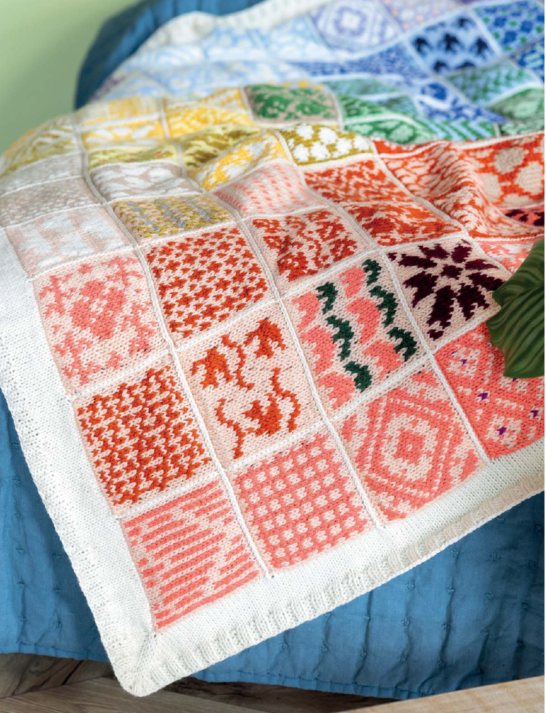 A close-up of a knitted patchwork blanket draped over a blue quilted bed. The blanket features multiple square panels with various colorful knit patterns in orange, red, pink, yellow, green, and blue on an off-white base.