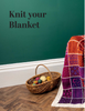 A styled promotional scene with a green wall, wood floor, a wicker basket holding colorful yarn balls, and a partially knitted patchwork blanket draped over the right side. Large text at the top reads 'Knit your Blanket'.