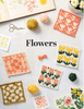 A styled flat-lay of knitted floral swatches arranged on a white surface with yarn balls, scissors, an open floral book, and small flower cards. The central text reads “Flowers.