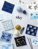 A flat lay of knitted sky-themed swatches in various blue, navy, yellow, and cream patterns, arranged with balls of yarn, knitting needles, scissors, and bird illustrations on a light background. The word 'Sky' is printed in the center.