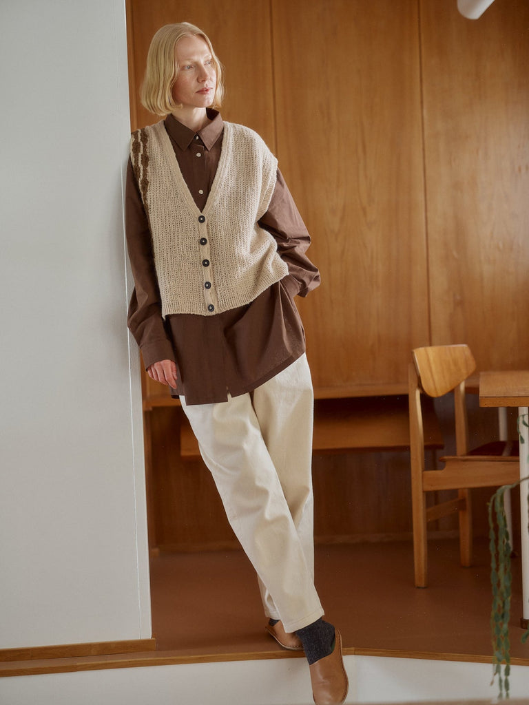 A woman models a brown button-up shirt layered under a cream-colored knit vest, paired with cream trousers and brown shoes, standing in a warm wood-paneled room.