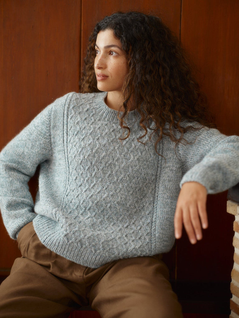 A woman with long curly dark hair wearing a light blue-gray cable-knit sweater and brown pants, seated against a warm brown wooden background.