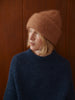 A woman wearing a warm rust-brown knit beanie and a dark navy ribbed sweater, standing against a dark wood-paneled background.