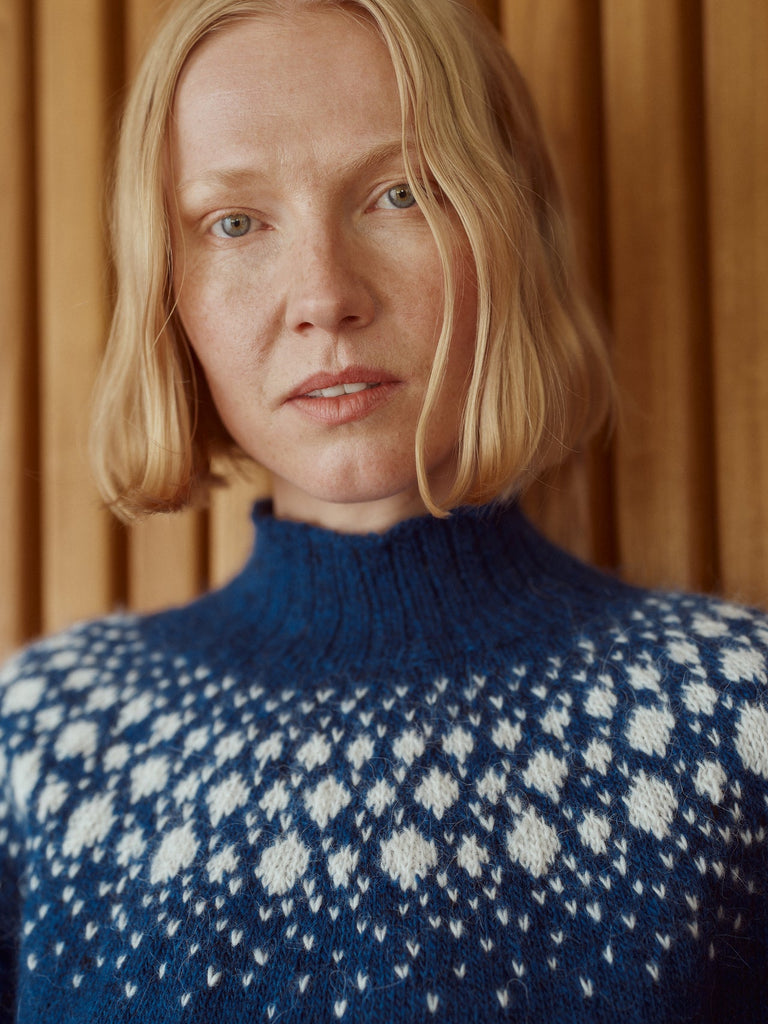 A close-up portrait of a woman with blonde hair wearing a blue and white patterned knit turtleneck sweater, set against a warm wooden background.