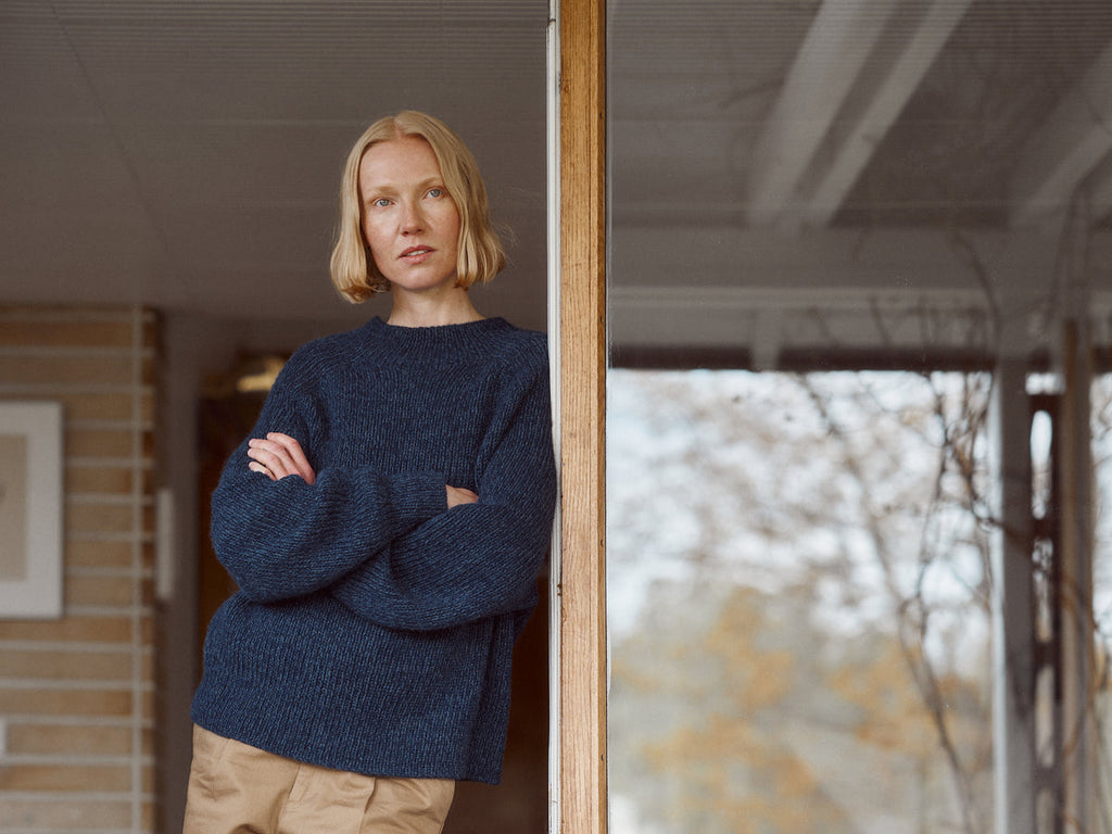 A woman with short blonde hair stands leaning against a wooden door frame, wearing a chunky navy blue knit sweater and tan pants, with a porch and blurred outdoor trees in the background.