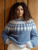 A woman with curly dark hair wearing a blue and white patterned knit sweater, standing against a wooden paneled wall.