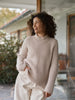 A woman with curly dark hair wearing a beige ribbed knit mock neck sweater and cream trousers, standing in front of a glass door of a house.