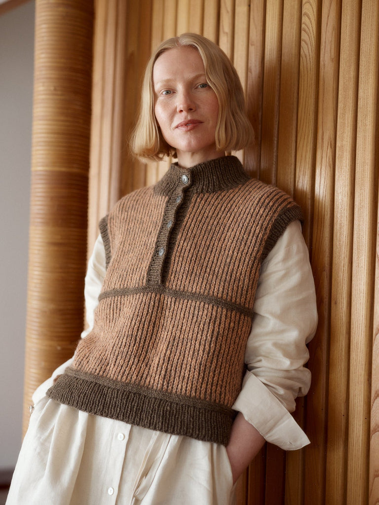 A woman with short blonde hair leans against a light wood slatted wall, wearing a brown ribbed knit sleeveless vest over a cream-colored long-sleeve shirt and white skirt.
