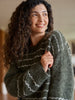 A smiling woman with long, curly brown hair wearing a fuzzy, oversized knit sweater in dark olive green with white horizontal stripes, photographed in a warm indoor setting.