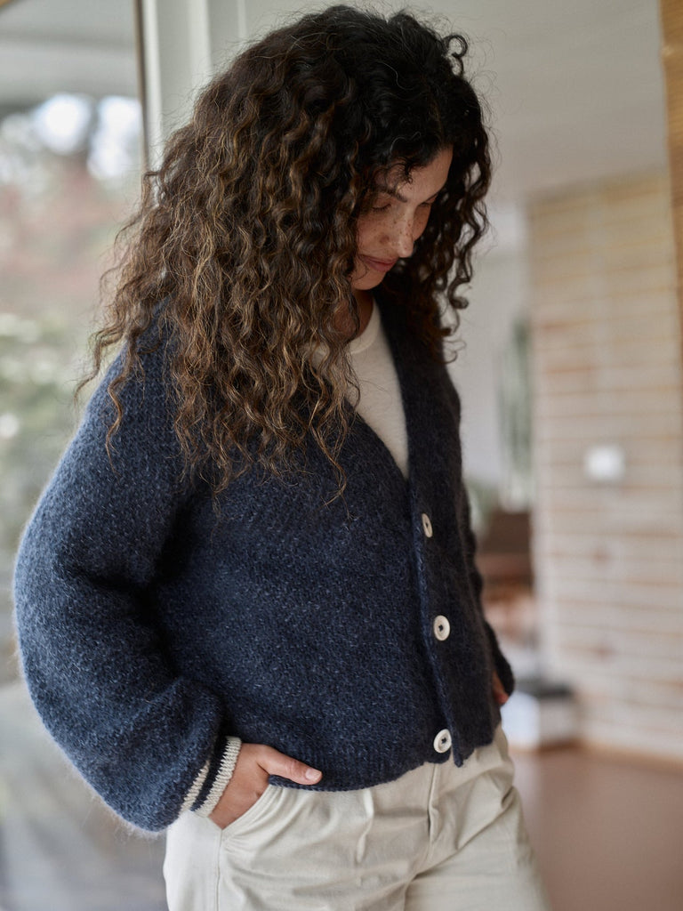 A woman with long, curly dark brown hair wearing a fuzzy navy blue cardigan over a cream top and light beige pants, standing in a softly lit indoor setting.
