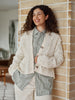 A woman with curly brown hair smiling and leaning against a beige brick wall, wearing a cream cable-knit cardigan over a green and white striped button-up shirt and cream pants.