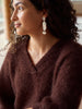 A close-up side profile of a woman with curly dark hair wearing a fuzzy brown V-neck sweater and a long, textured white dangle earring.