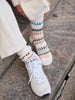 A close-up of a person sitting on stone steps, wearing cream knit socks with multicolor geometric patterns and white sneakers.