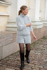 A woman wearing a light gray fuzzy knit mock-neck sweater and matching shorts, styled with black tights and black leather knee-high boots, standing on a cobblestone street in front of a cream-colored building.