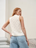 A woman wearing a cream cable-knit sleeveless vest and light blue jeans, photographed from behind in an outdoor setting with a light-colored wall and stone steps.