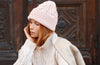 A woman wearing a light pink cable-knit beanie and a cream cable-knit sweater, standing in front of a dark brown wooden door.
