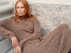 A woman with red hair wearing a chunky cable-knit turtleneck sweater and matching skirt in a warm taupe-brown color, sitting on stone steps.