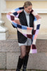 A woman wearing a black blazer, gray turtleneck, and black knee-high boots, holding up a chunky fuzzy plaid scarf in pastel pink, blue, purple, beige, and white.